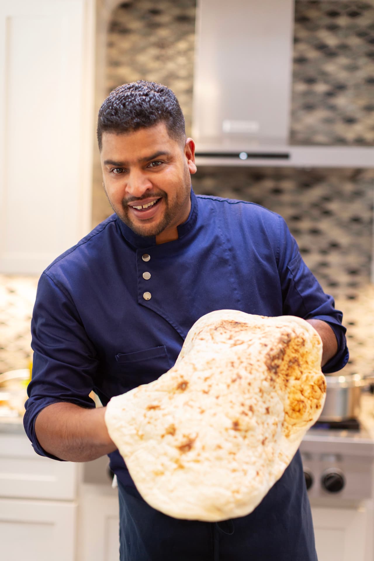 Chef Youssef Hsaini smiling in his kitchen, holding up a large piece of freshly grilled pita bread